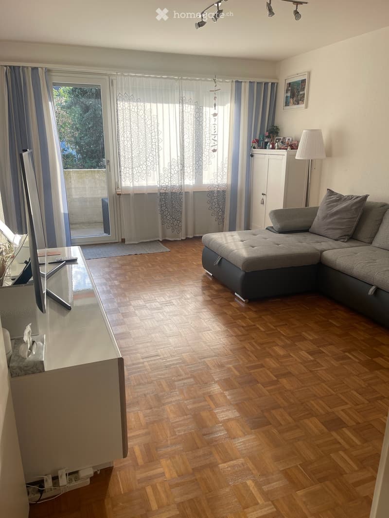 Living room with wooden parquet flooring, grey sectional sofa, white side table, floor lamp, and large window with curtains leading to a balcony.