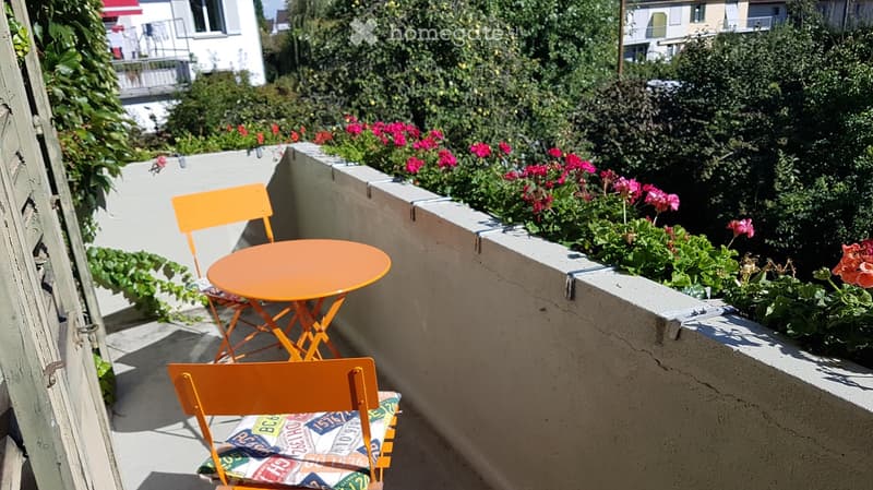 Small balcony with orange table and chairs, surrounded by colorful flowers and greenery, overlooking neighboring buildings.