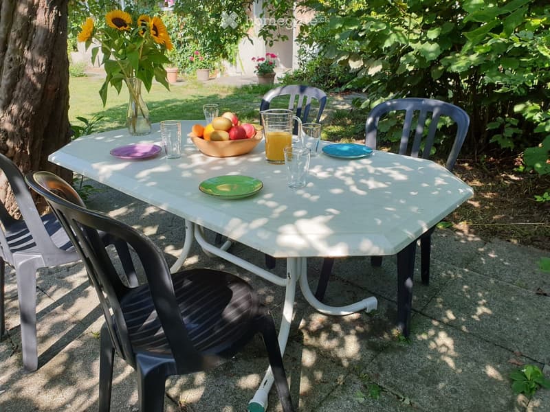 A garden patio with a white table, surrounded by plastic chairs, a vase with sunflowers, a bowl of fruit, and several glasses and plates.