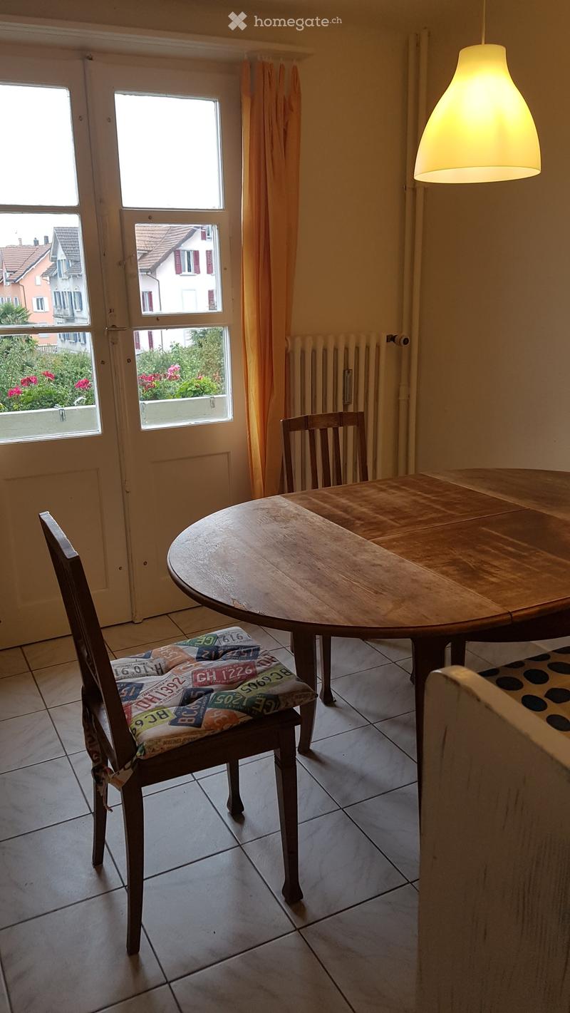 A small, well-lit room with a round wooden table and a single chair with a colorful cushion. The room has tiled flooring, a radiator, and large windows with orange curtains, offering a view of a garden and neighboring buildings.
