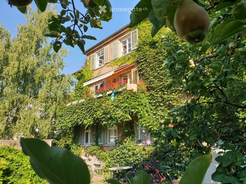 A two-story house covered in lush greenery, with a balcony adorned with flowers and a small outdoor seating area with a table and chairs.