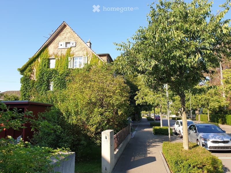A two-story house with ivy-covered walls, multiple windows, and a chimney, situated on a tree-lined street with parked cars and greenery.
