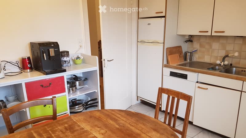 Modern kitchen with white cabinets, a refrigerator, dishwasher, sink, coffee maker, and a small dining area with a wooden table and chairs.