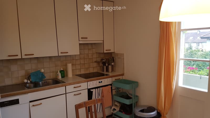 The kitchen features white cabinets, a sink with a faucet, an oven, and a dishwasher. There is a small green rolling cart with various items, a wooden chair, and a window with an orange curtain overlooking a garden.
