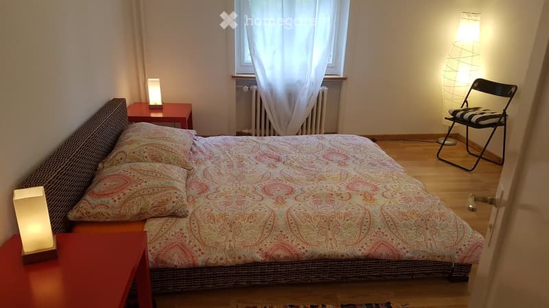 Cozy bedroom with a double bed, patterned bedspread, two nightstands with lamps, a wooden floor, and a folding chair near a window with white curtains.