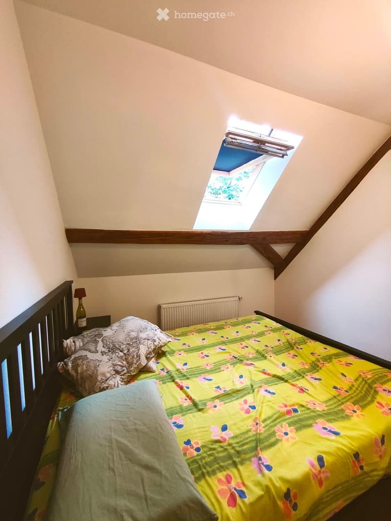 Cozy attic bedroom with a single bed, colorful floral bedspread, nightstand with lamp, and skylight window above the bed.