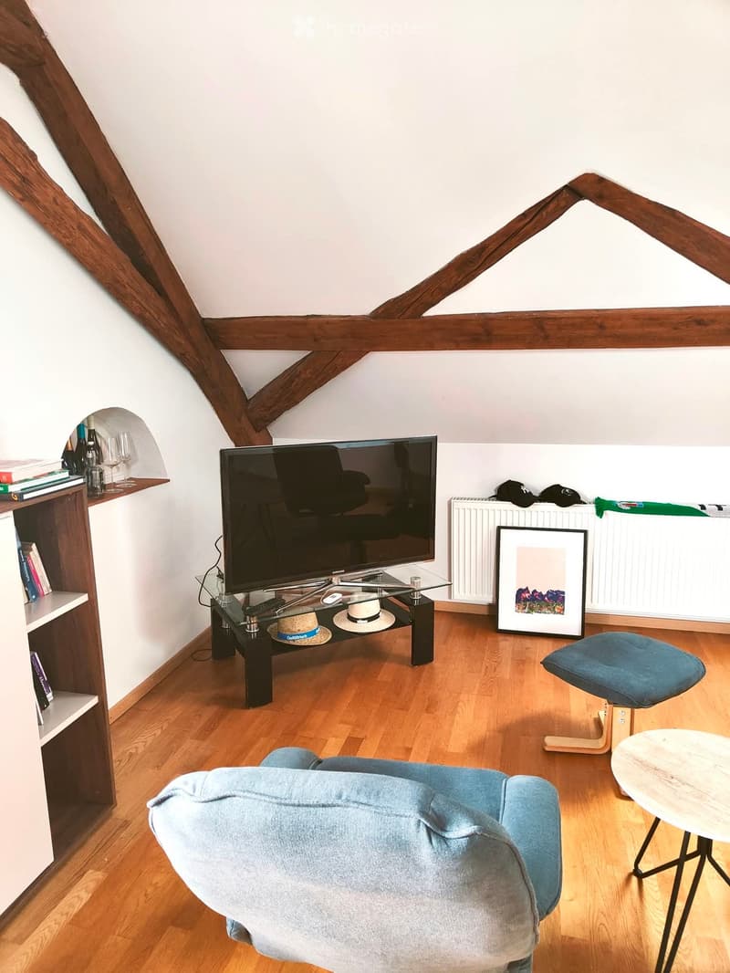 Living room with wooden floors, exposed wooden beams, a blue sofa, a glass coffee table with a TV, a bookshelf, and a small round table.