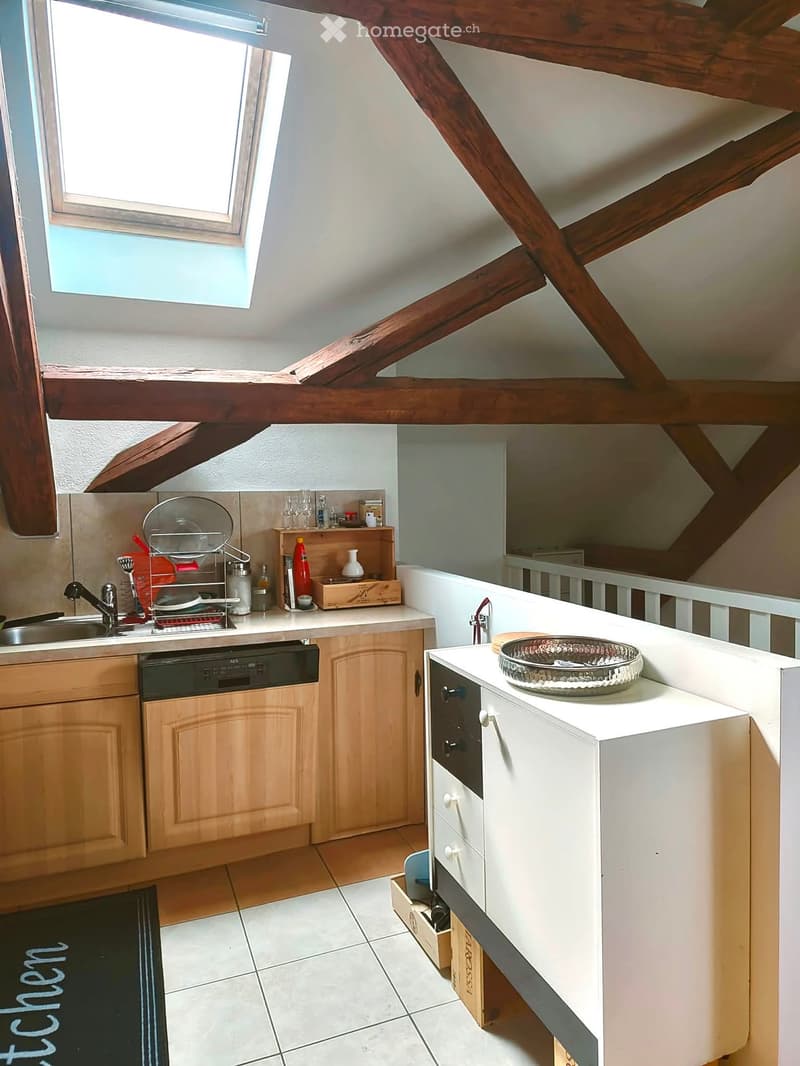 A compact kitchen with wooden cabinets, a white countertop, a sink, a dishwasher, and a small bowl on the counter. The space features exposed wooden beams and a skylight window.