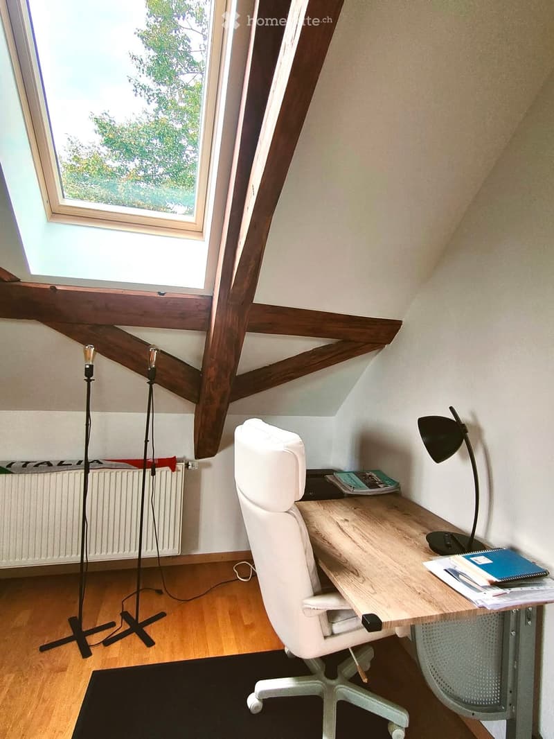 Bright attic room with wooden beams, large skylight, white leather chair, wooden desk, floor lamp, and books.