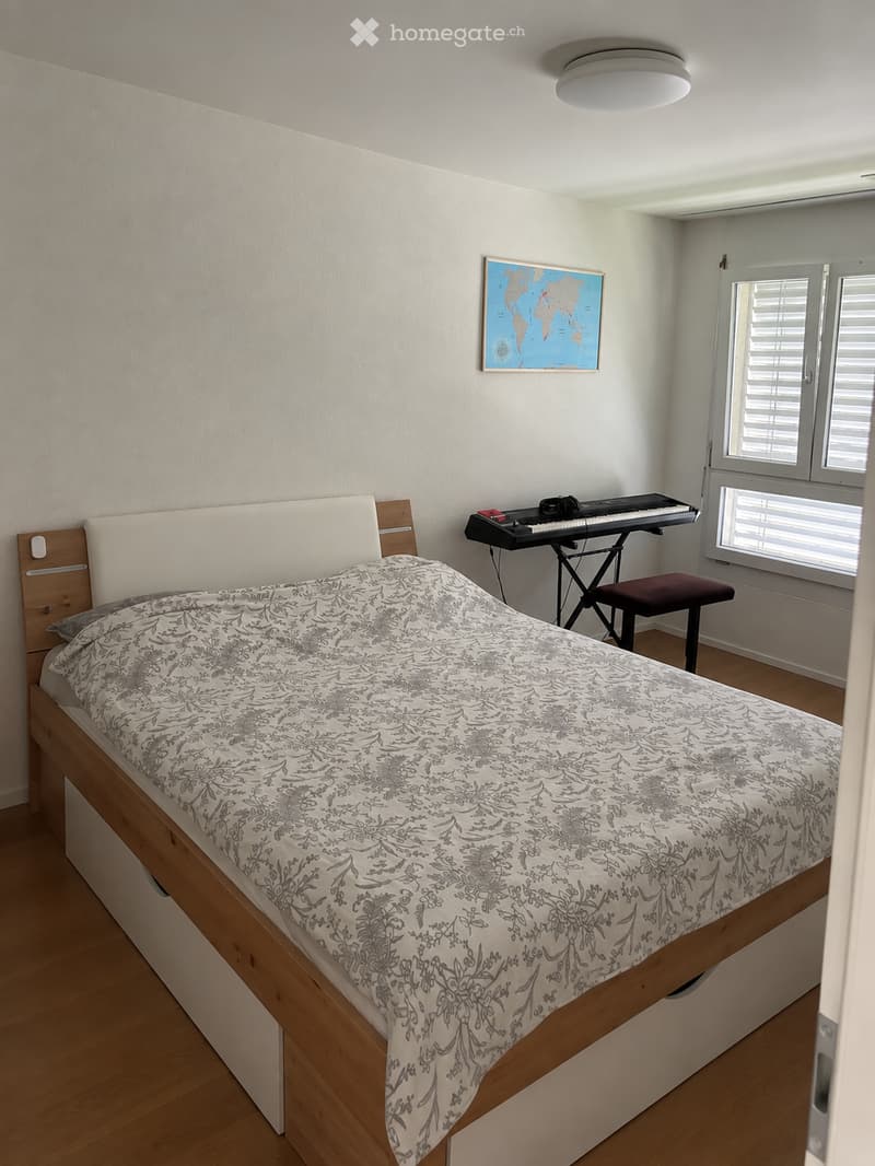 A small, well-lit bedroom with a wooden bed, patterned bedding, a keyboard on a stand, and a world map on the wall.