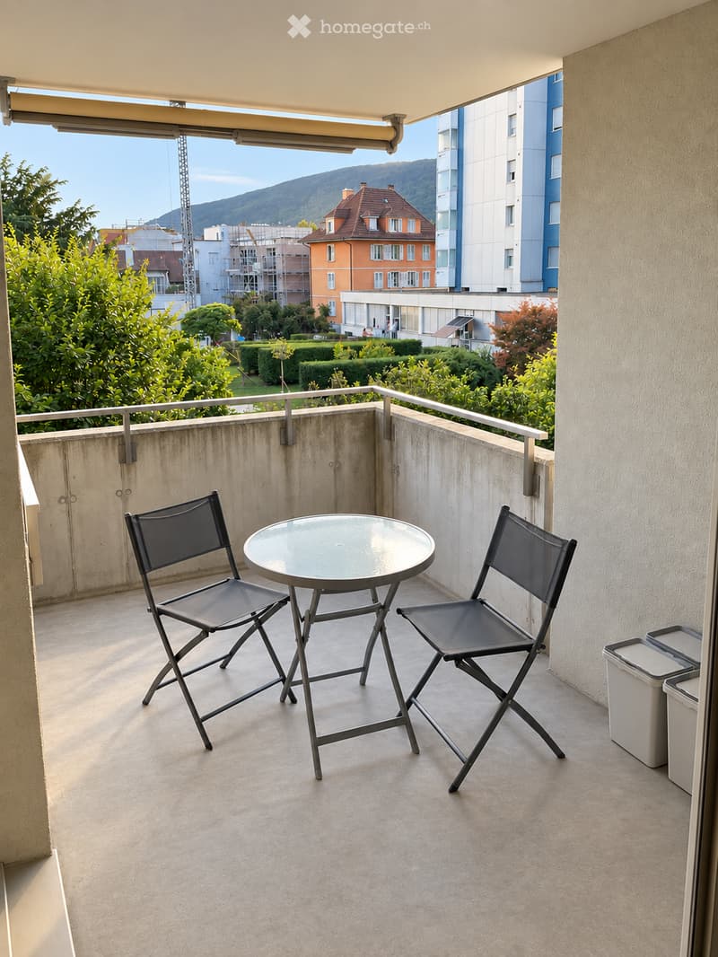 Balcony with a round glass table and two folding chairs, overlooking a landscaped area with trees and buildings.