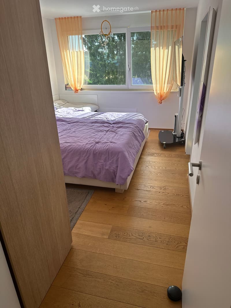 Bedroom with wooden flooring, a bed with purple bedding, a small table with a screen, and orange curtains on the windows.