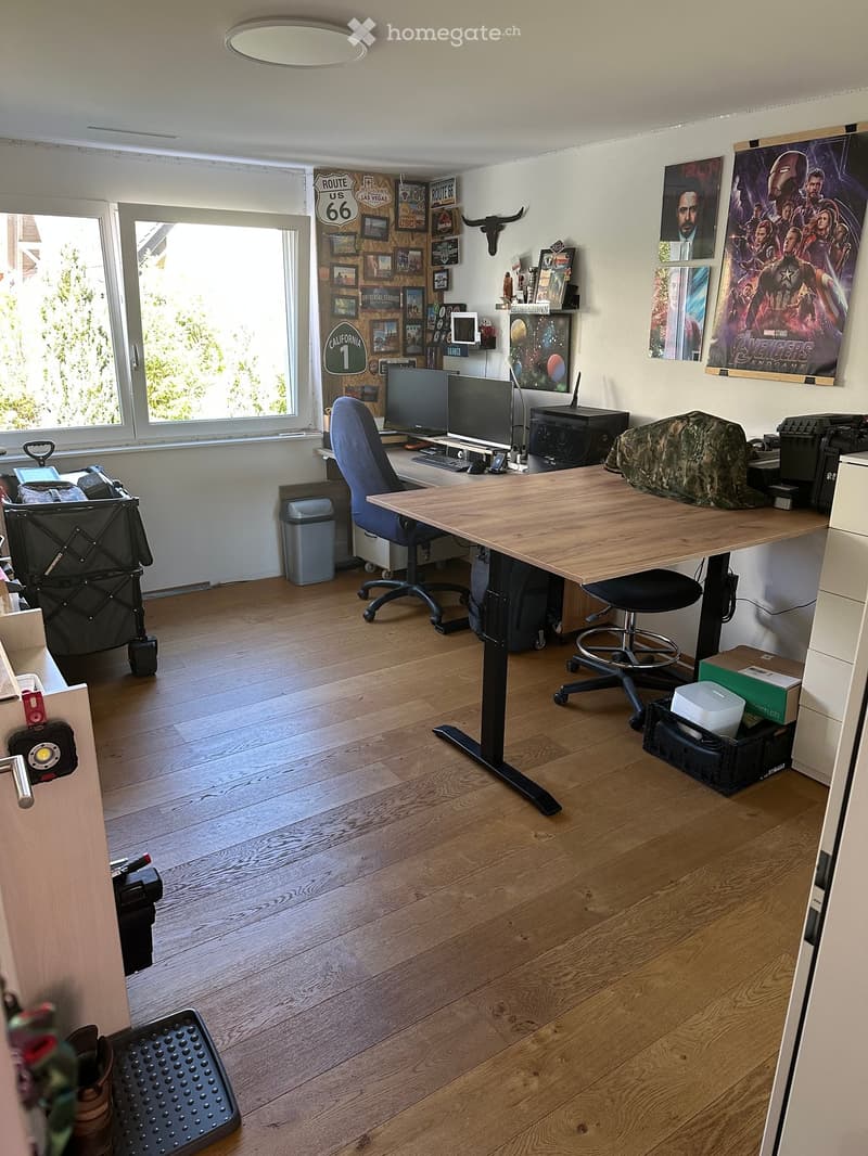 A well-lit home office with a standing desk, ergonomic chair, computer setup, and various decorative items including posters and framed pictures. The room features wooden flooring and large windows allowing natural light.