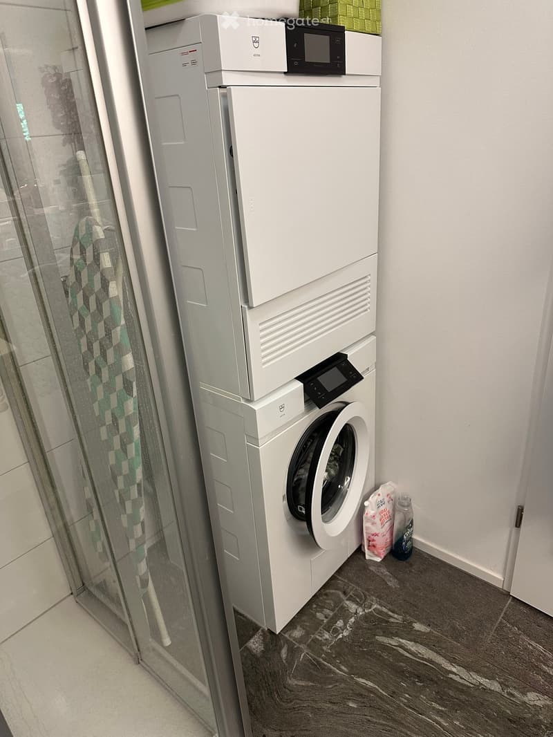 Modern bathroom with a stacked washing machine and dishwasher unit, dark tiled floor, and a glass shower enclosure with a green and white patterned curtain.