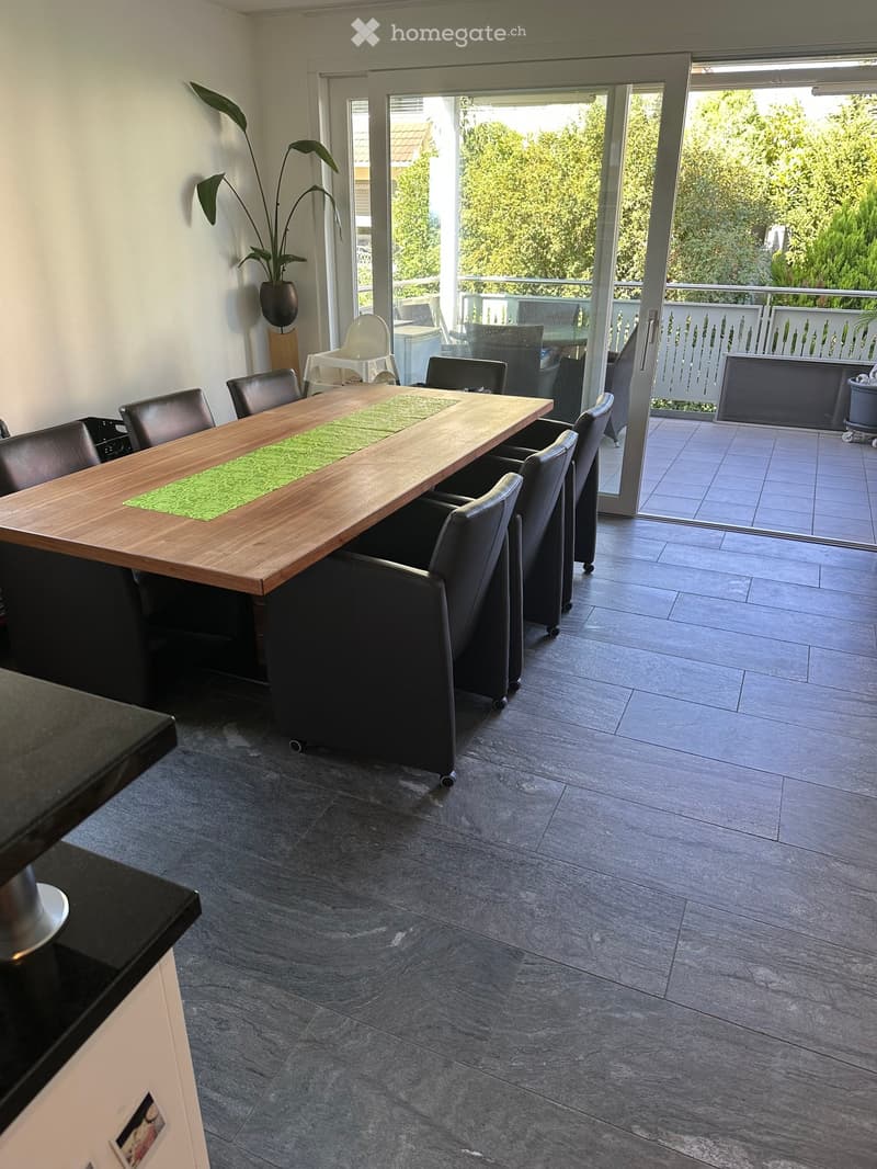 Dining room with a long wooden dining table, black chairs, and a green placemat. A large sliding glass door leads to a balcony with outdoor furniture.