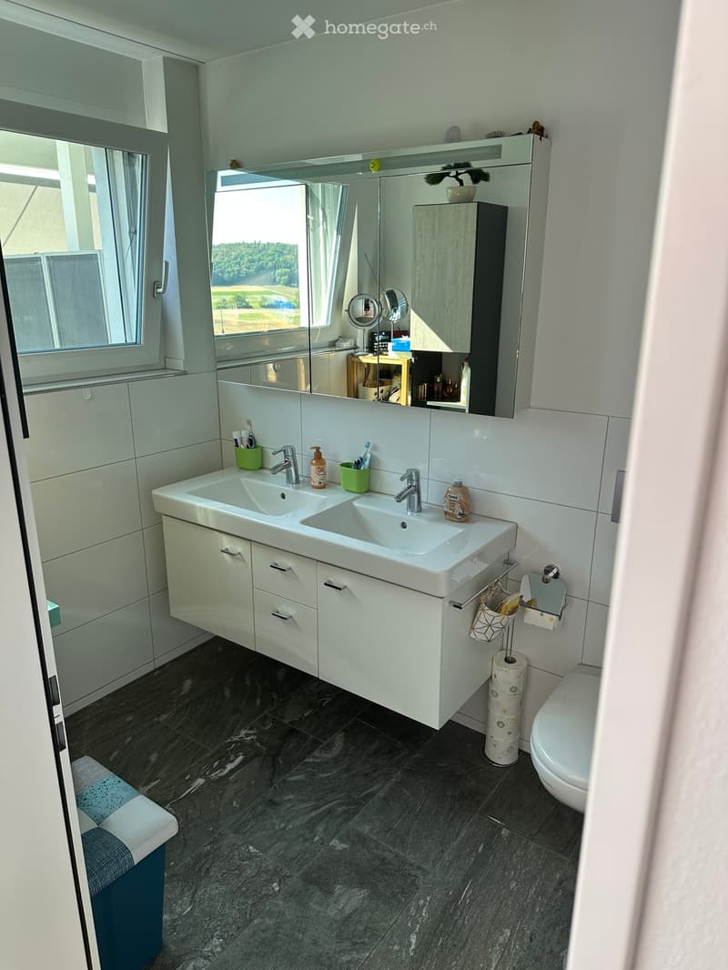Modern bathroom with double sink, large mirror, white cabinets, and a view of greenery through the window.