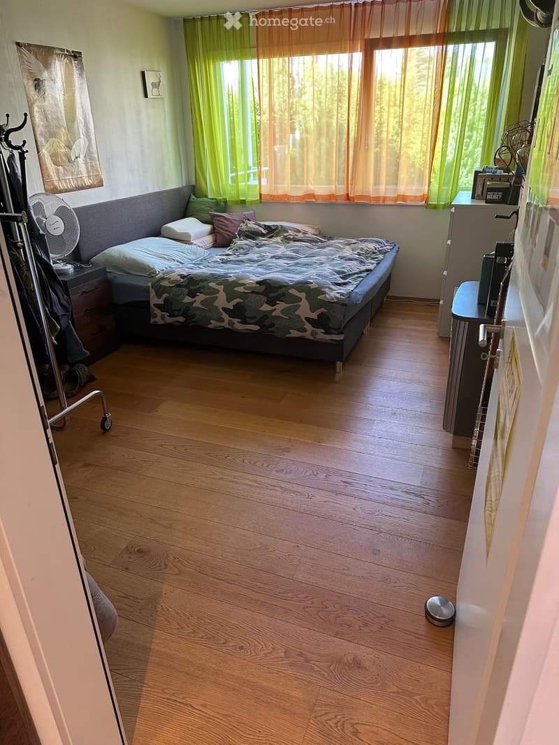 Cozy bedroom with wooden flooring, a bed with a camouflage-patterned blanket, nightstands, a fan, and a curtained window.