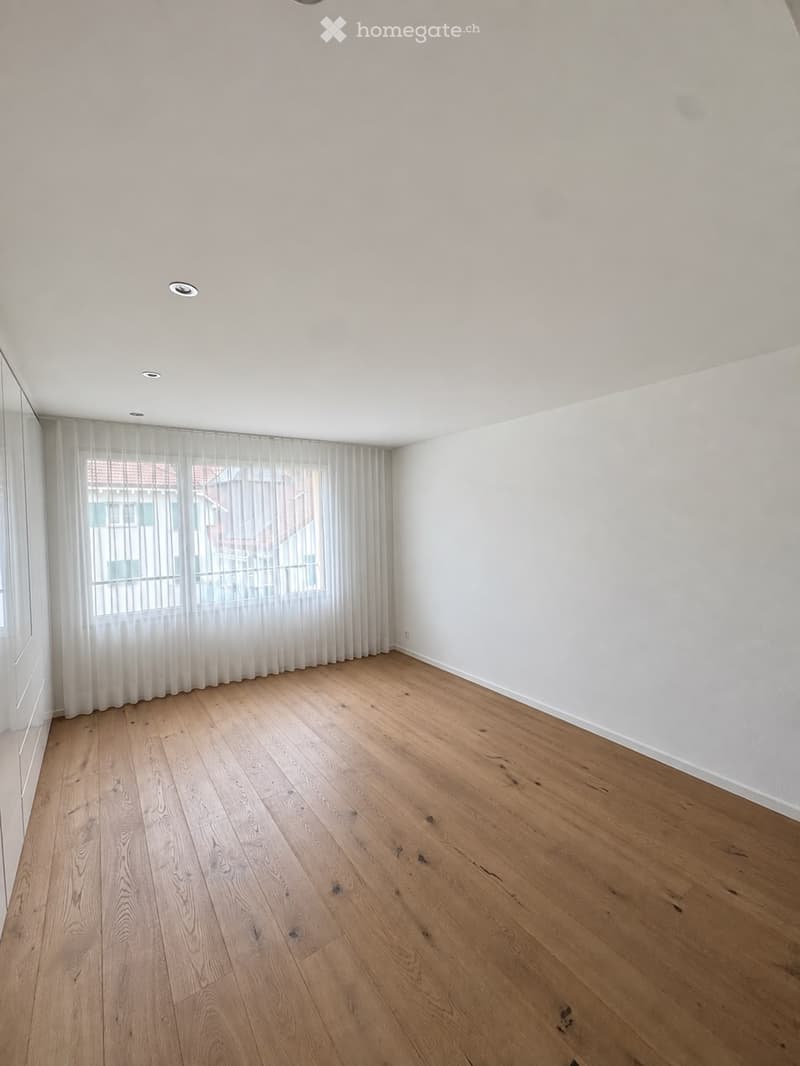 Empty room with light wooden flooring, white walls, and large windows covered with sheer curtains. The room has recessed ceiling lights and a visible door frame on the left side.