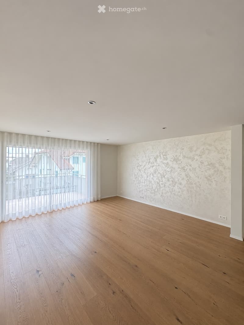 Empty room with light wooden flooring, white walls, and large windows with white curtains. The room has a modern design with recessed ceiling lights and a decorative textured wall.