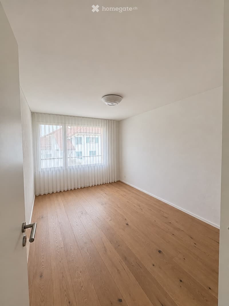 Empty room with light wooden flooring, white walls, and a large window with sheer curtains. A modern ceiling light fixture is visible.