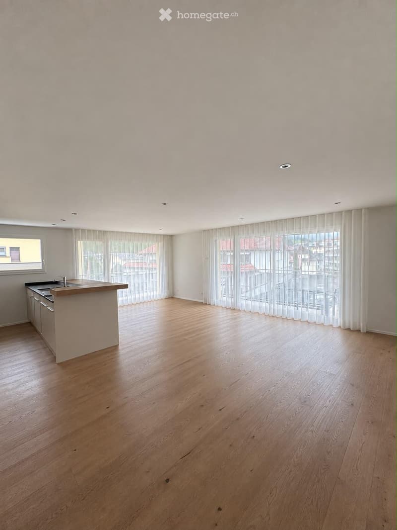 Empty room with light wooden flooring, white walls, and large windows with sheer curtains. A small kitchen area with cabinets and a countertop is visible on the left side.