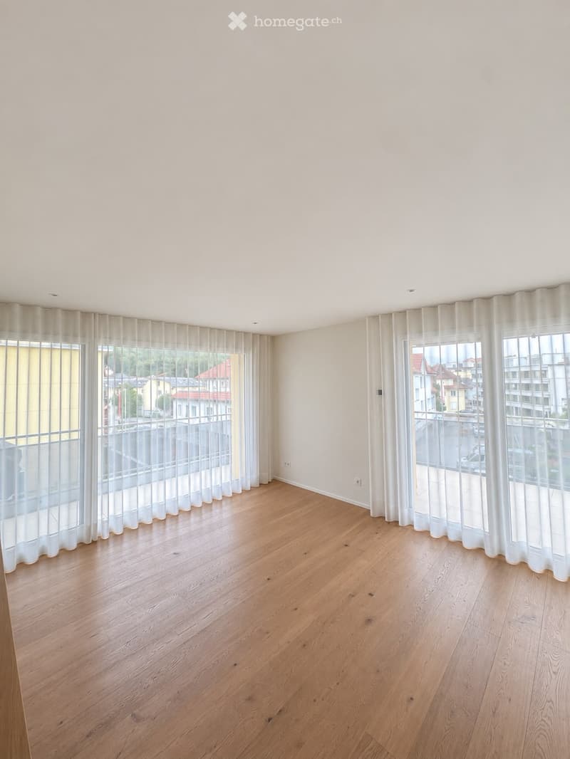 Empty room with wooden flooring, large windows with sheer curtains, and a view of the street and buildings outside.