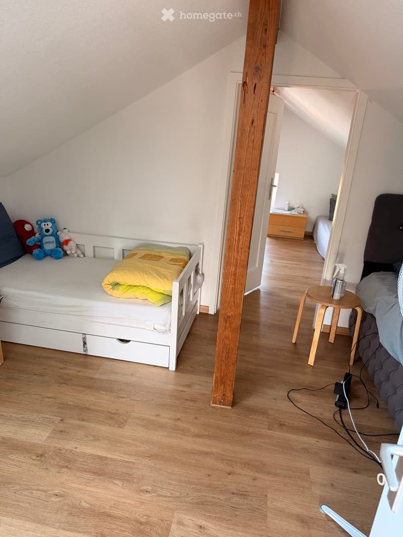 Cozy attic bedroom with wooden flooring, a single bed with a yellow blanket, a small wooden stool, and a fan. The room features slanted ceilings and a visible wooden beam.