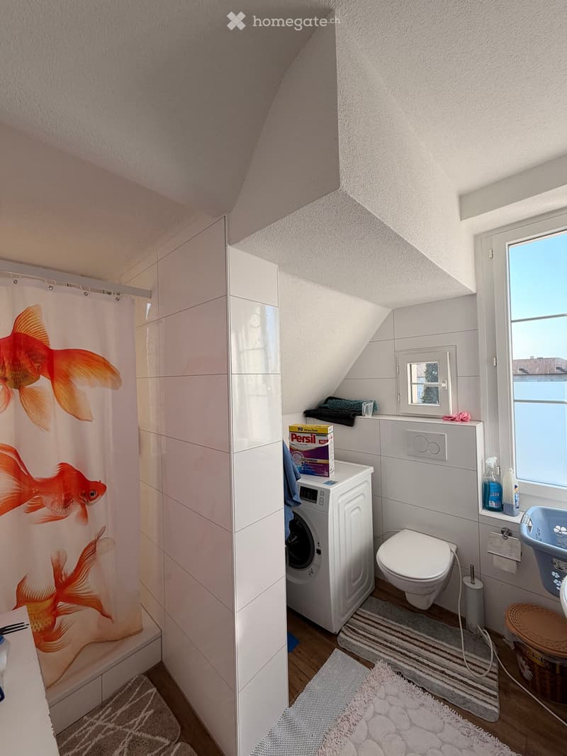 Bathroom with a toilet, washing machine, shower curtain, sink, and various toiletries. The room features a slanted ceiling and a window with a small opening.