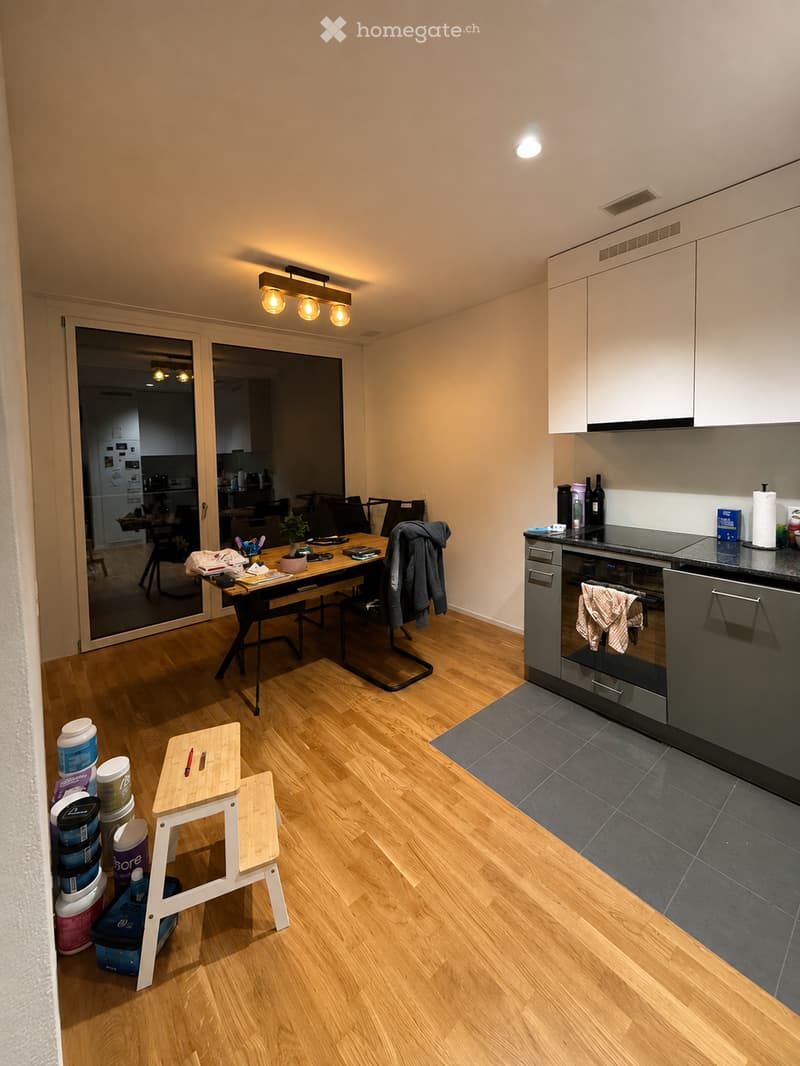 The image shows a modern, well-lit kitchen and dining area with light wooden flooring, white cabinets, and a black countertop. A small wooden stool is placed near the entrance, and a dining table with chairs is visible in the adjacent space. The kitchen f