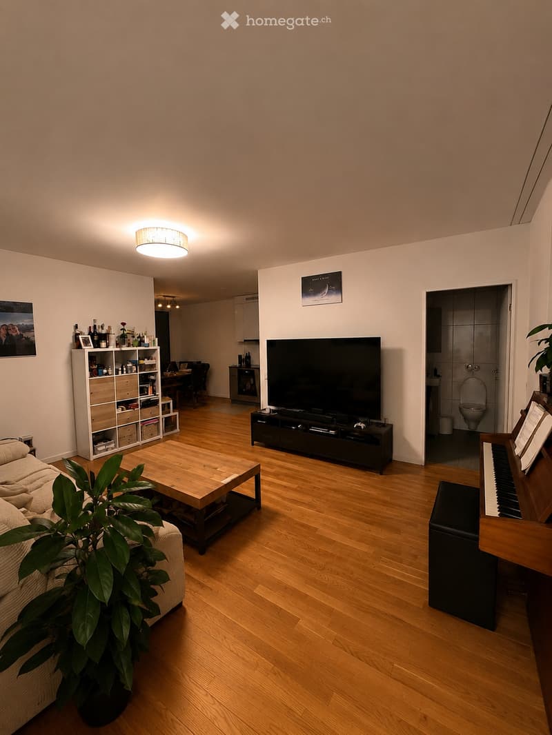 Living room with wooden flooring, a beige sofa, a wooden coffee table, a TV stand with a flat-screen TV, a piano, a storage unit, and a dining area visible in the background.