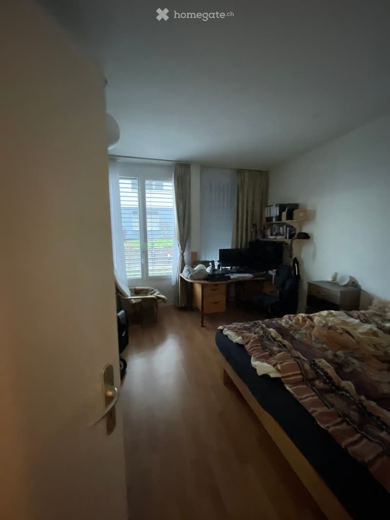 Well-lit bedroom with wooden flooring, a bed with patterned bedding, a desk with a computer, a chair, and a window with curtains.