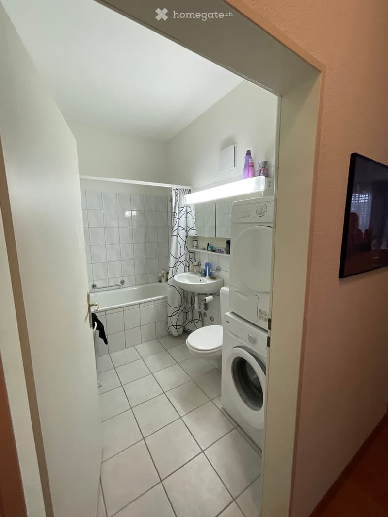 Bathroom with a bathtub, toilet, sink, washing machine, and a mirror. The room has tiled walls and floor, and a curtain in the shower area.