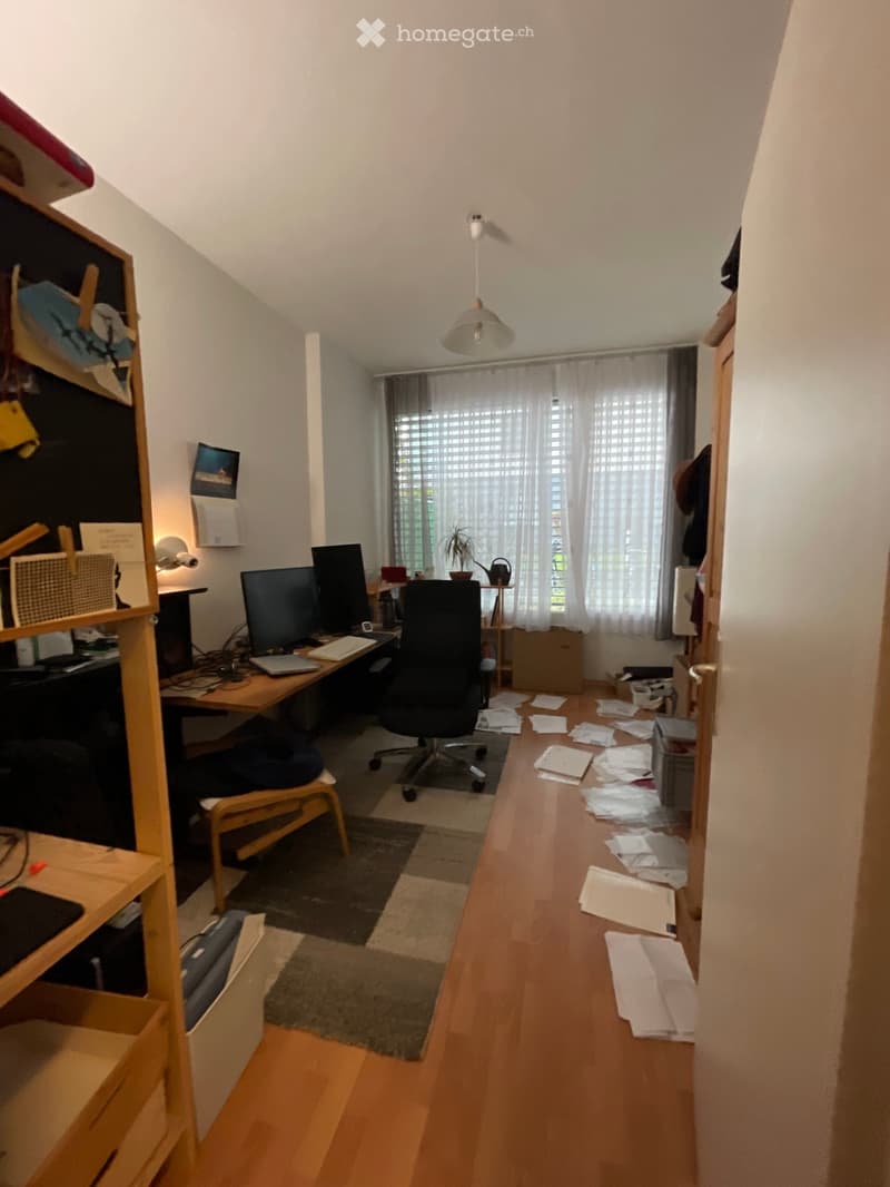A well-lit study or home office with a wooden desk, ergonomic chair, laptop, dual monitors, keyboard, and a small shelf with books and office supplies. The room features wooden flooring, a large window with blinds, and a small plant on a side table.