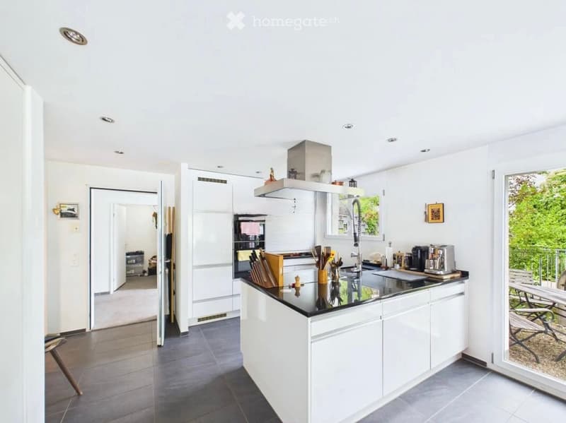 Modern kitchen with white cabinets, black countertop, stainless steel appliances, and a large island. Features a coffee maker, utensils, and a view of a balcony area.