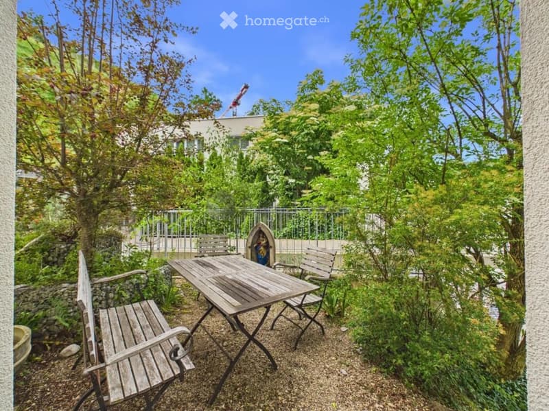 A small, enclosed garden area with a wooden table and two chairs, surrounded by lush greenery and trees, with a view of a building in the background.