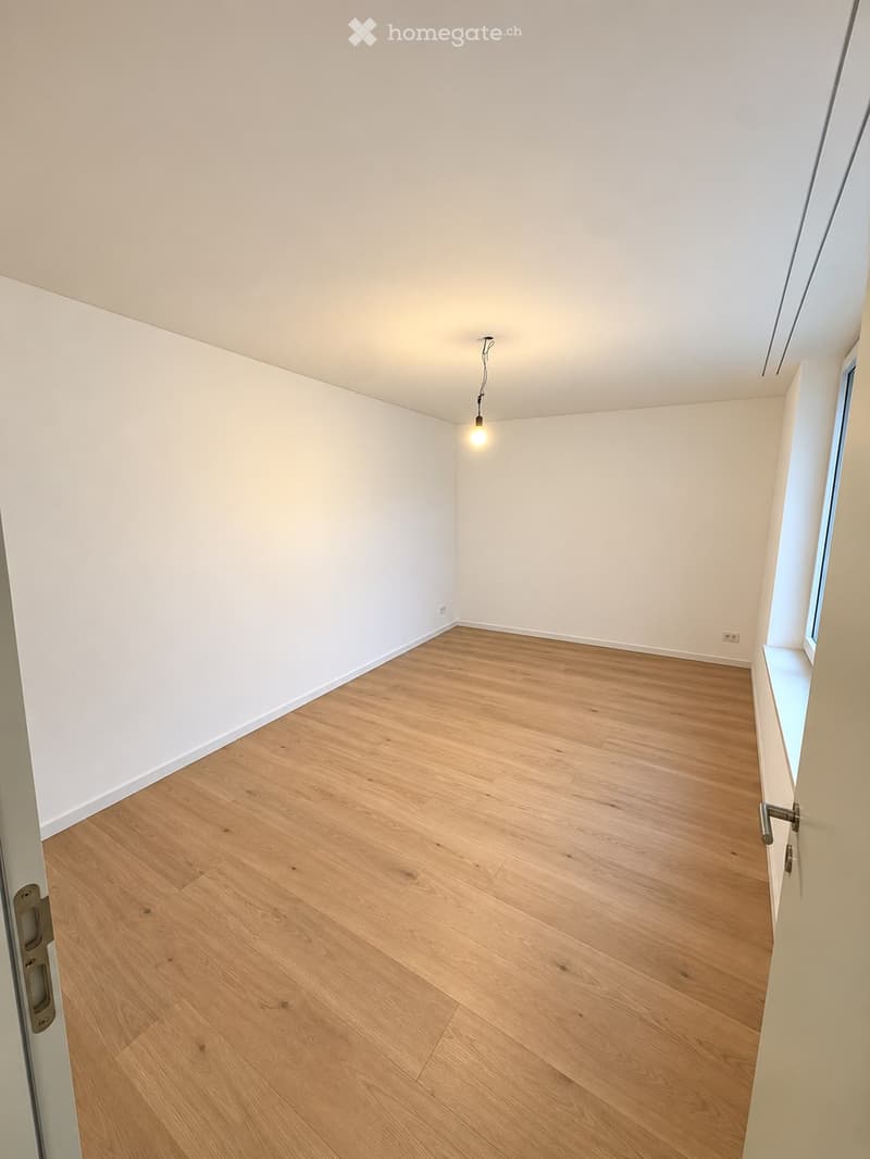 Empty room with light wooden flooring and white walls, featuring a single light fixture and a small window on the right side.