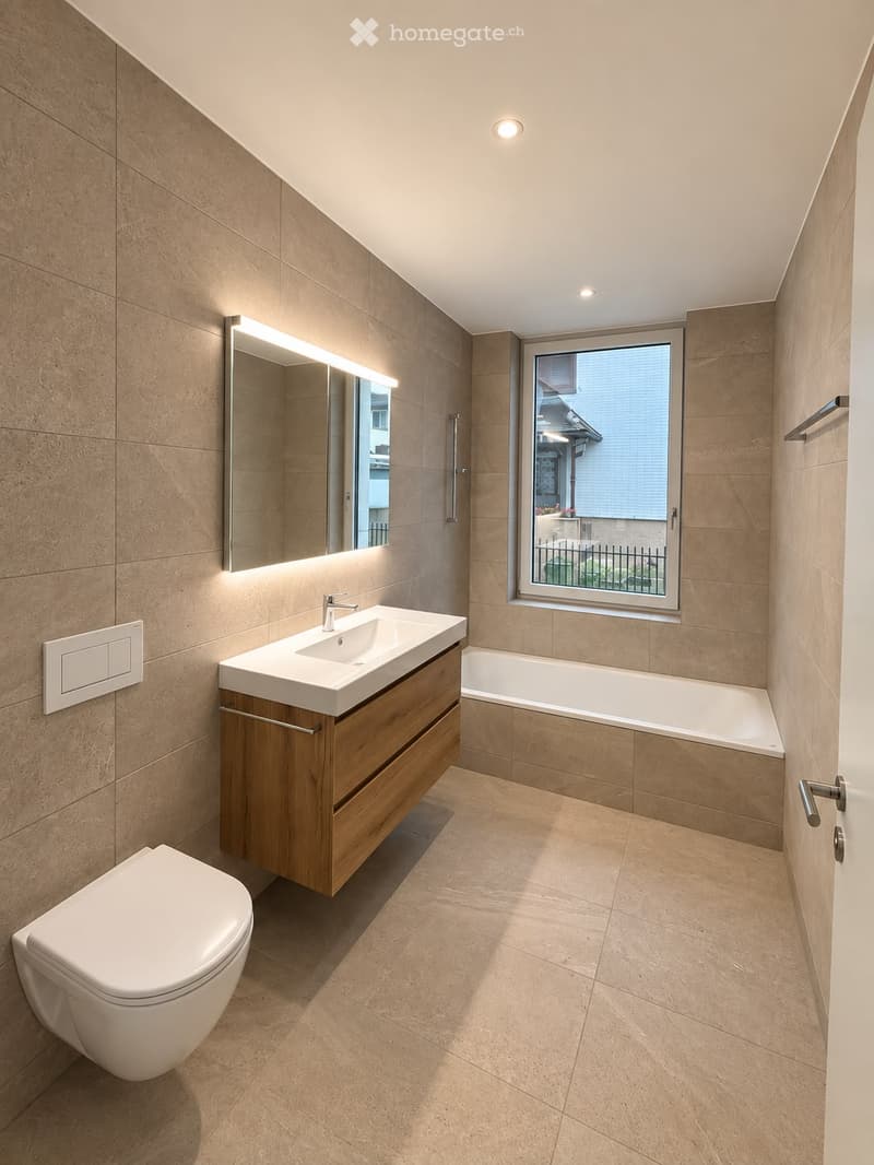 Modern bathroom with beige tiles, a white sink with wooden cabinet, a toilet, a bathtub, and a large mirror with LED lighting. A window offers a view of the outside.