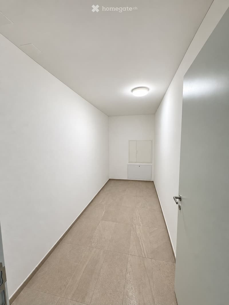 Empty hallway with white walls, beige tiled floor, and a single ceiling light fixture. A small cabinet is visible at the end of the hallway.