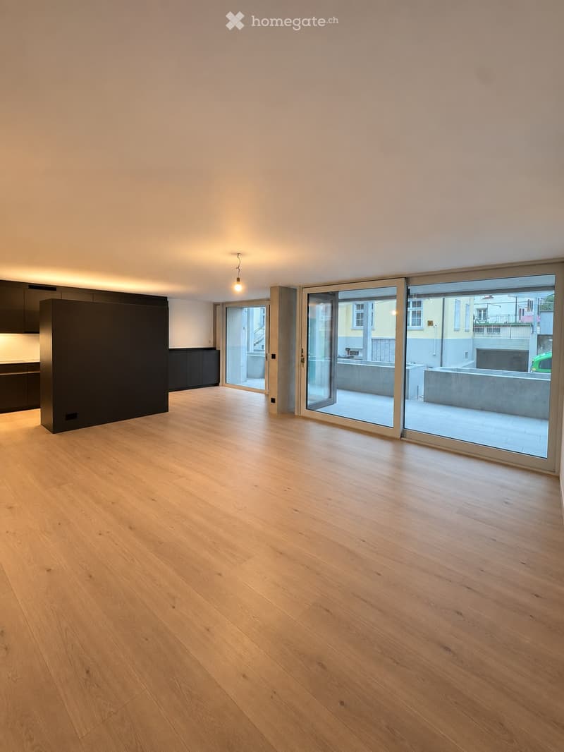 Empty room with light wooden flooring, large sliding glass doors leading to a balcony, and a modern kitchen area visible in the background.