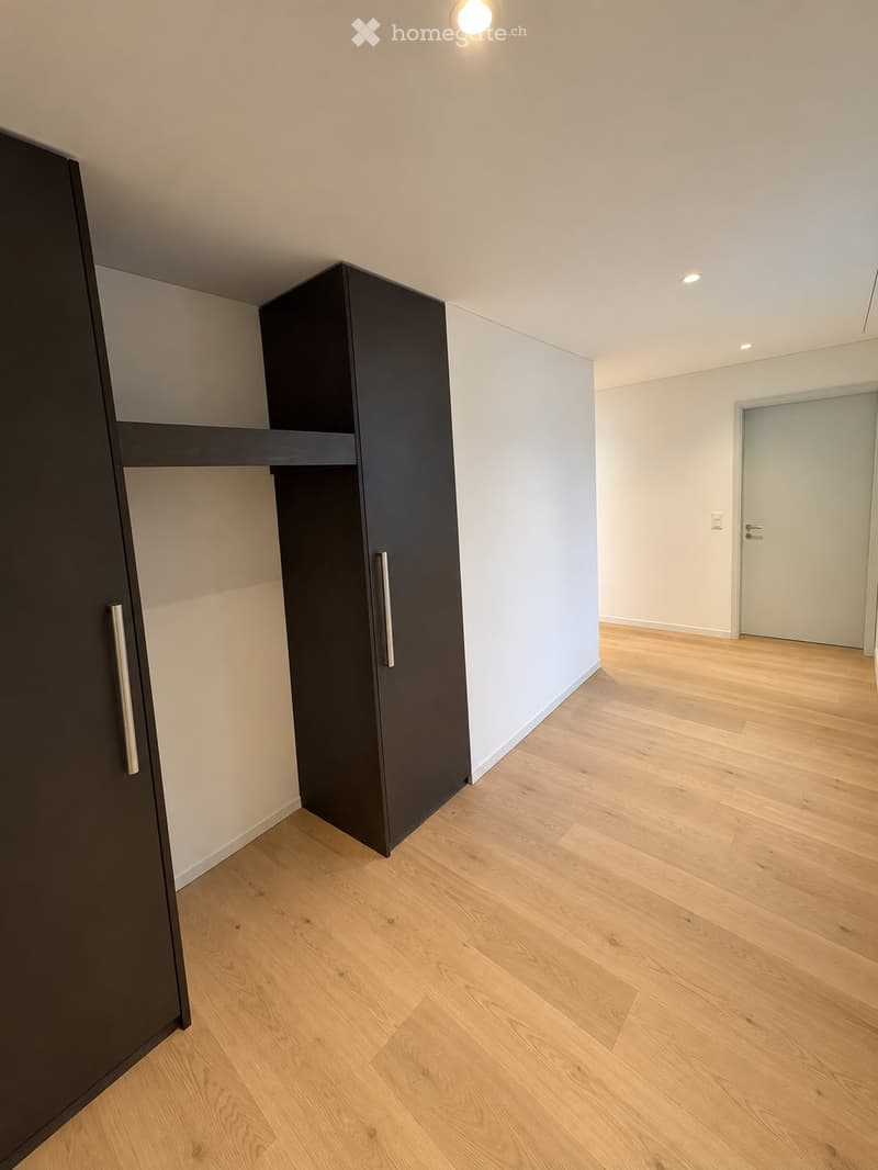 Empty room with light wooden flooring, white walls, and built-in dark-colored closet units. A closed door is visible in the background.