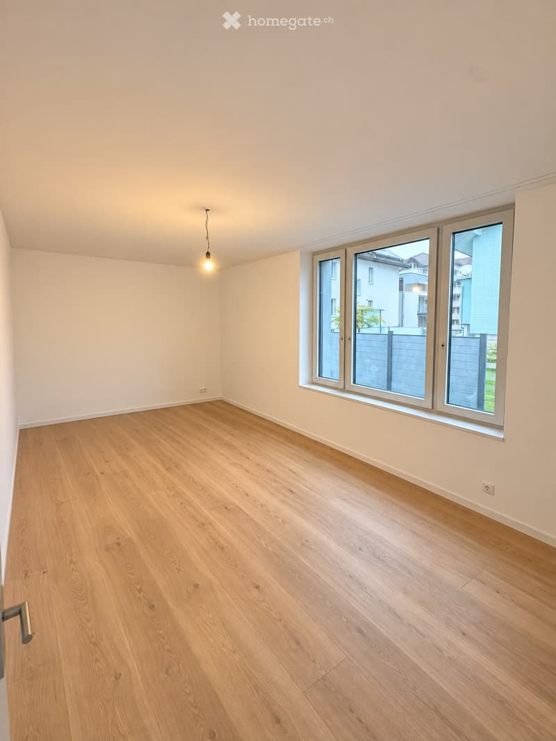 Empty room with light wooden flooring, white walls, and large windows offering a view of the outside. A single pendant light hangs from the ceiling.