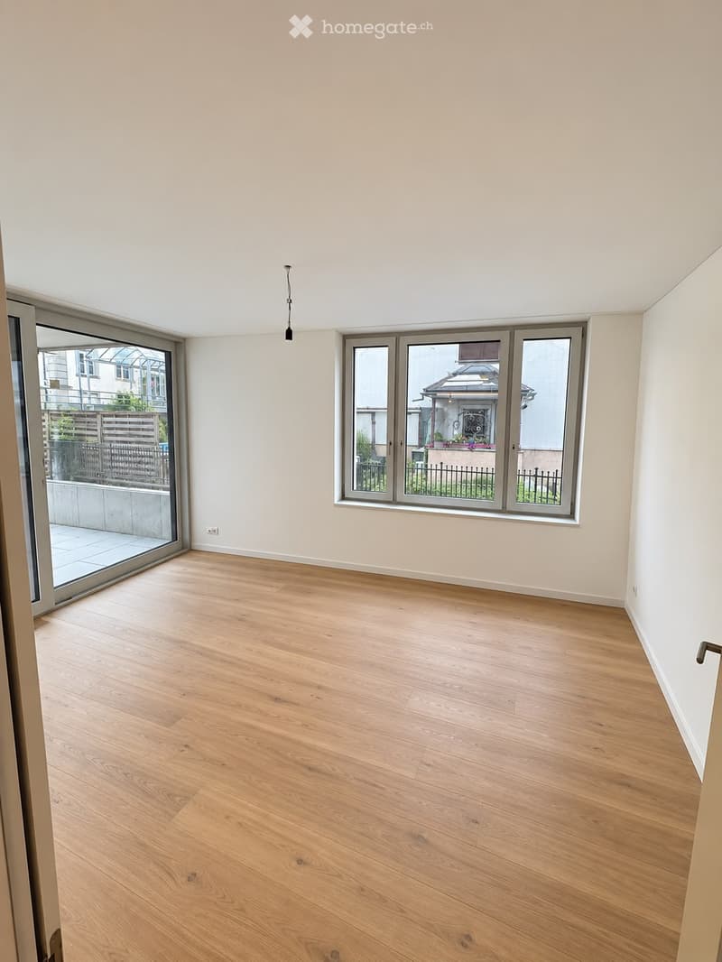 Empty room with light wooden flooring, white walls, large windows, and a sliding glass door leading to a balcony area.