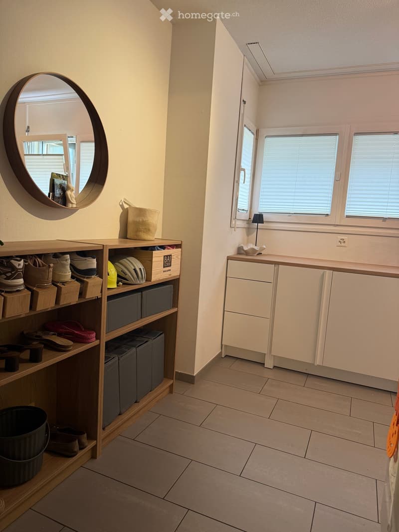 A well-lit hallway with tiled flooring, white walls, and built-in storage units. Features include a round wall mirror, a small shelf with shoes and storage boxes, and a cabinet with drawers and doors.