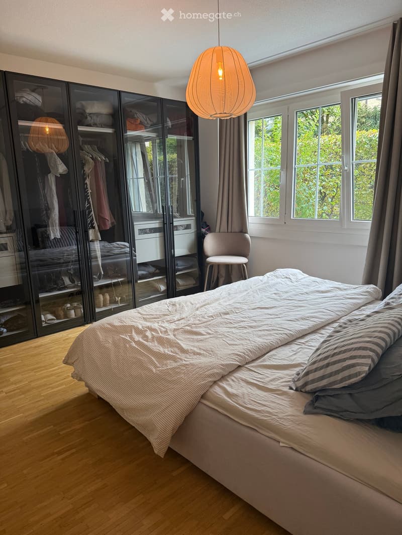 Modern bedroom with a large bed, striped bedding, a sleek black wardrobe with mirrored doors, a hanging pendant light, and a small chair near the window.