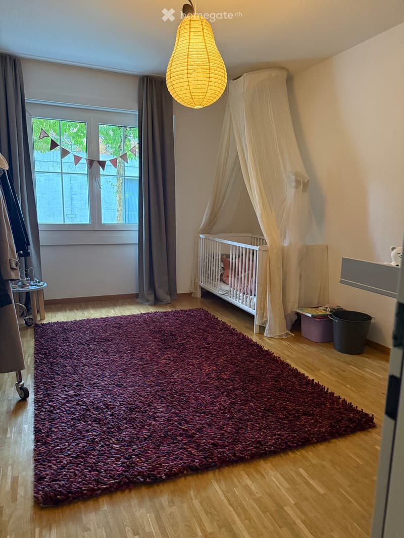 Brightly lit bedroom with wooden flooring, large window with curtains, crib with a canopy, plush red shag rug, small table, and decorative items.
