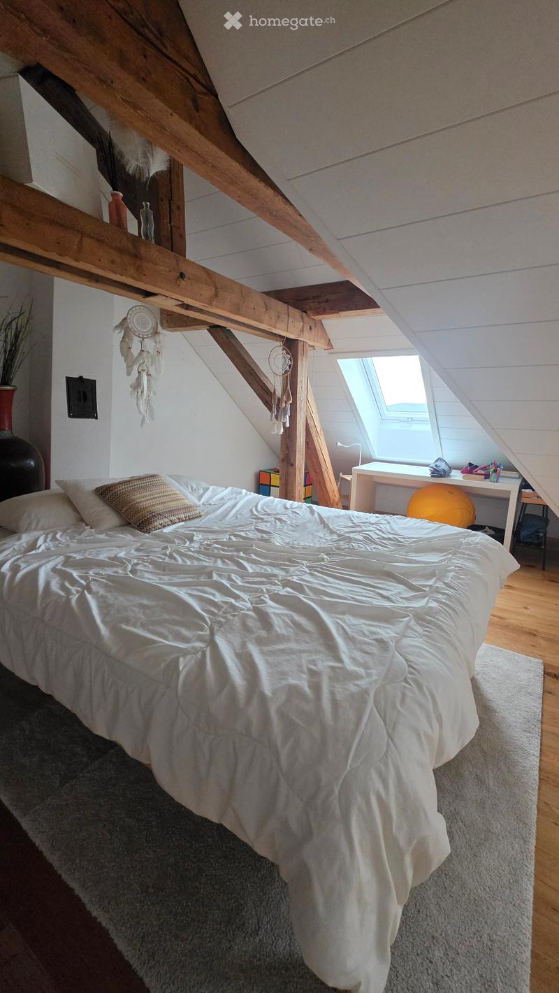 Cozy attic bedroom with wooden beams, a large bed with white bedding, a small desk with a chair, and a large window letting in natural light.
