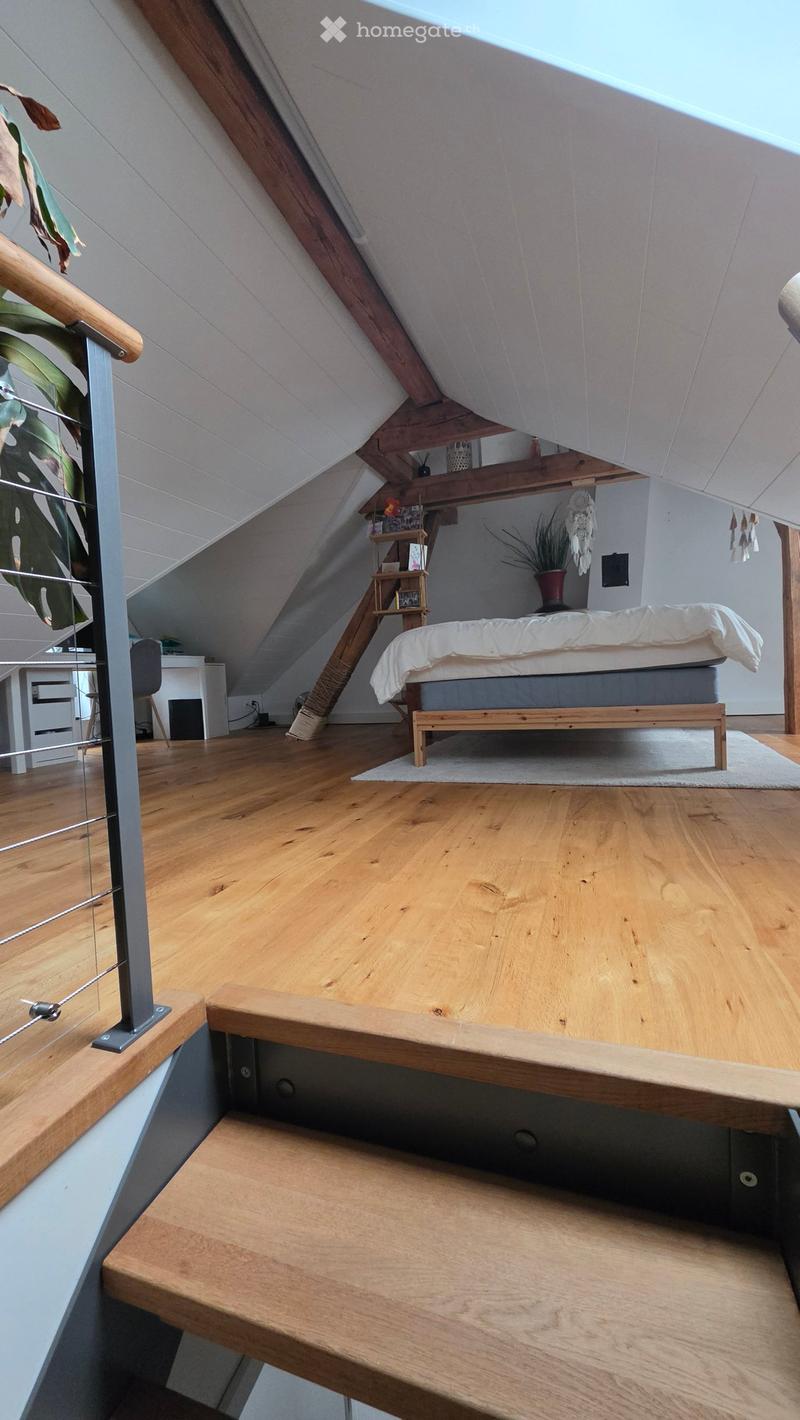 Attic bedroom with wooden floors, slanted ceilings, a bed with white bedding, a small table with a plant, a chair, and a cat tree.