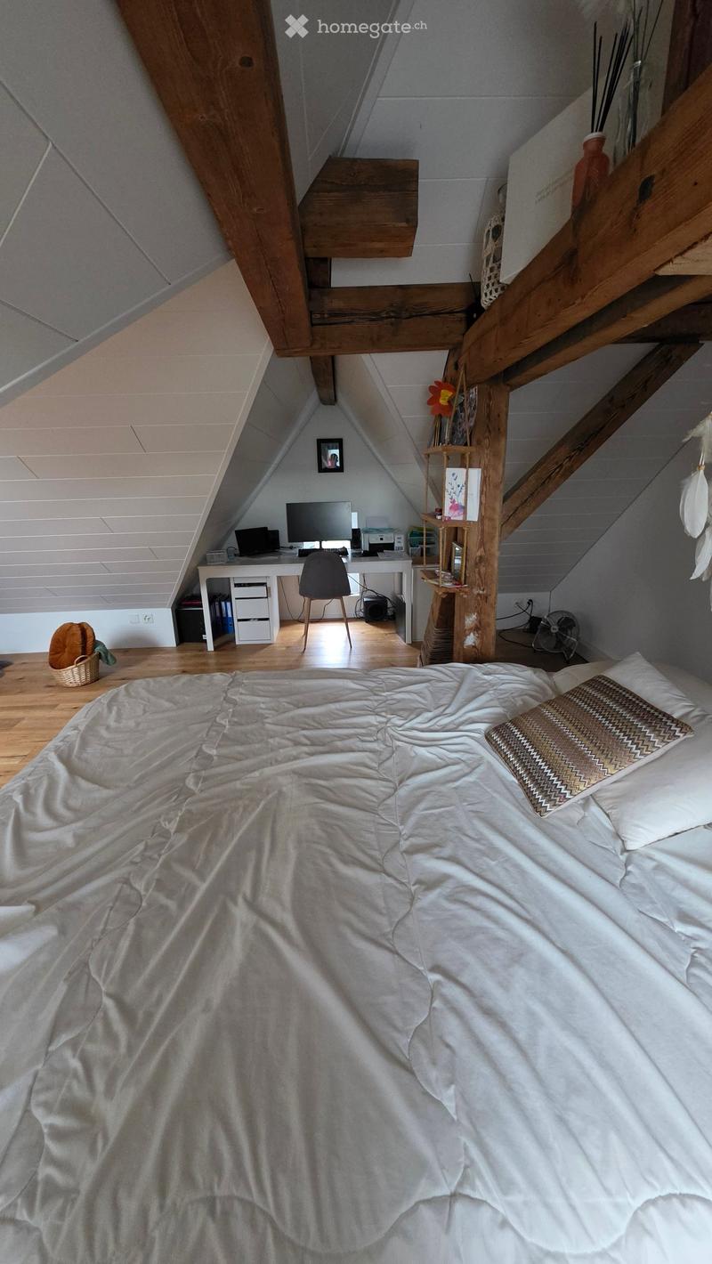 Cozy attic bedroom with a large bed, wooden floor, slanted ceilings, and a small desk area with a laptop and chair. Includes a shelf with books and decorative items, a fan, and a small basket on the floor.