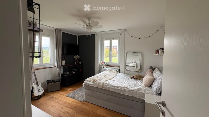 Cozy bedroom with wooden flooring, a large bed with pillows and a blanket, a mirror, a guitar, a speaker, shelves, a table with a vase, and a radiator. The room has large windows with curtains and a fan on the ceiling.