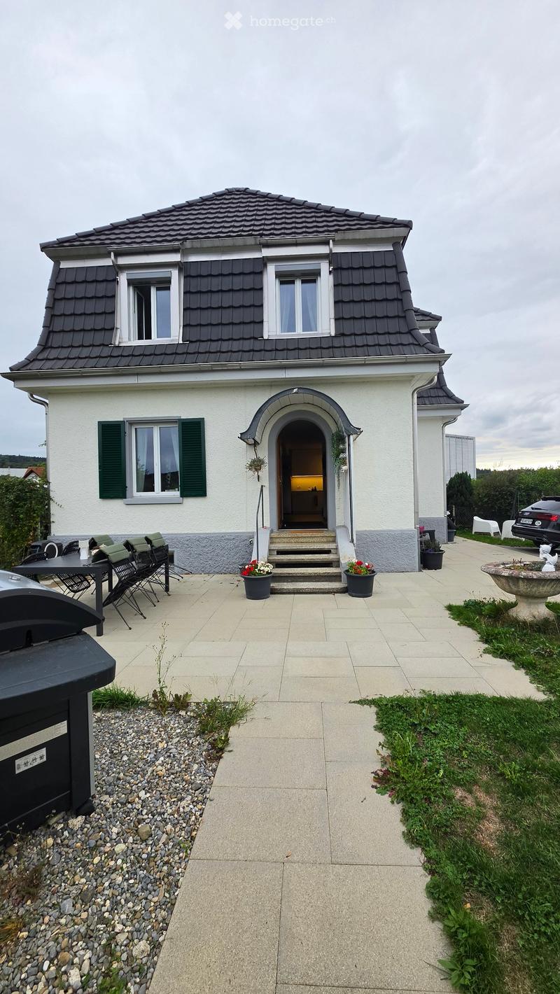 Two-story house with a gray roof, white walls, green shutters, and a small patio area with a table and chairs.